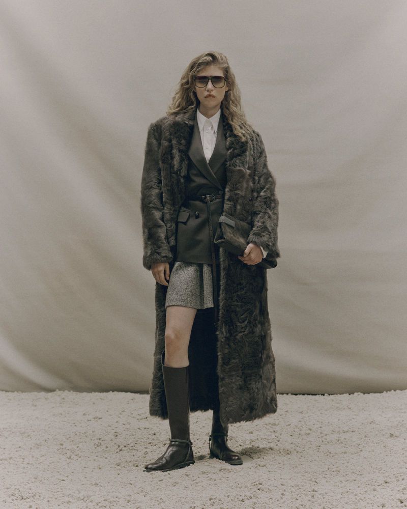 A stylish woman with wavy hair stands in a studio, wearing sunglasses, a long fur coat, knee-high socks, and boots, with a neutral background.