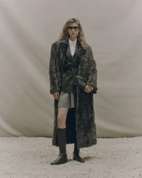Model wearing a floor-length dark textured fur coat, tailored blazer, mini skirt, sunglasses, and knee-high leather boots.