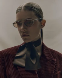 Model Mariam de Vinzelle wearing oval wire sunglasses, a black neck scarf, and a burgundy corduroy blazer against a neutral studio backdrop.