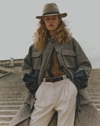 Model in a grey tweed coat, beige fedora hat, and white pleated trousers standing on outdoor stone steps.