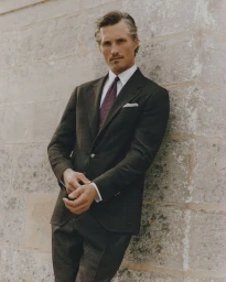 Ollie Edwards leaning against a stone wall wearing a dark tailored plaid suit, crisp white shirt, and burgundy necktie.