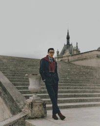 Man wearing a dark suit with a draped red sweater and glasses standing against large stone steps and classical architecture.