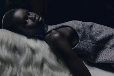 Model reclining on white fur wearing a grey herringbone textured dress and beaded choker in low-key lighting.