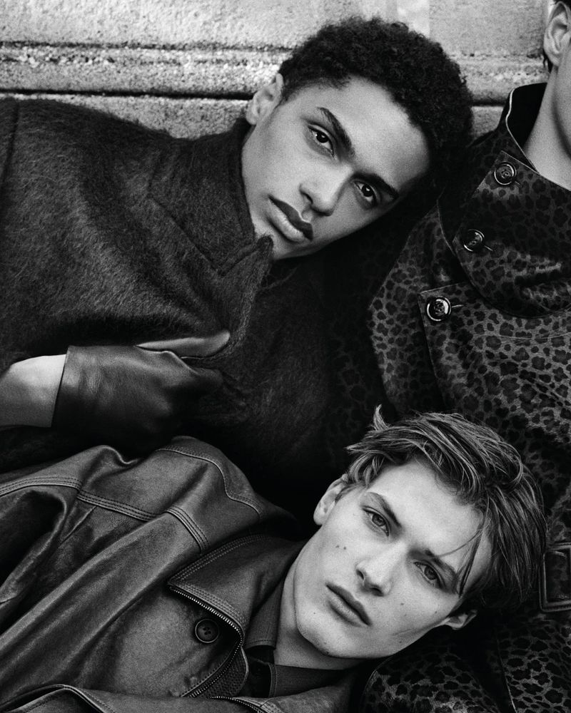 Three young men in leather and patterned coats are posing indoors, in a monochromatic, high-fashion studio-style portrait.