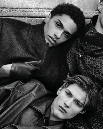 Three young men in leather and patterned coats are posing indoors, in a monochromatic, high-fashion studio-style portrait.