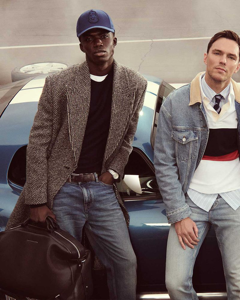 Two men pose next to a blue car. One wears a cap and coat, carrying a bag, while the other has a denim jacket and tie.