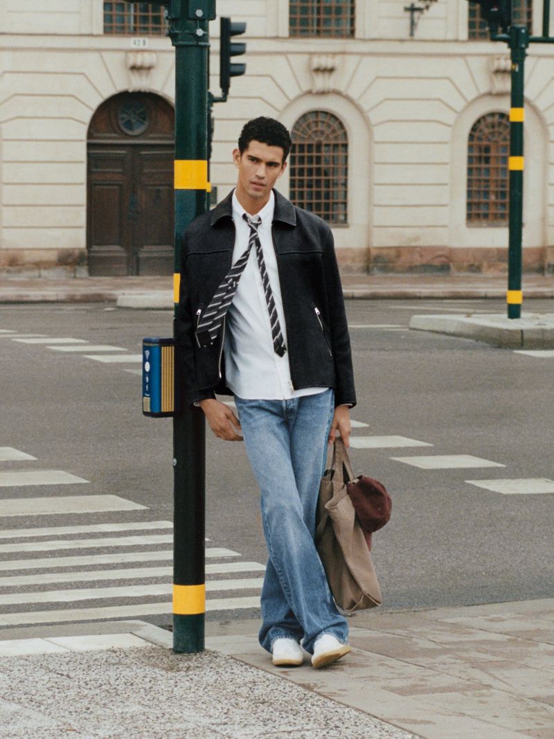 Man in jacket and jeans stands on urban crosswalk with traffic light, holding a bag, in daylight with architectural backdrop.