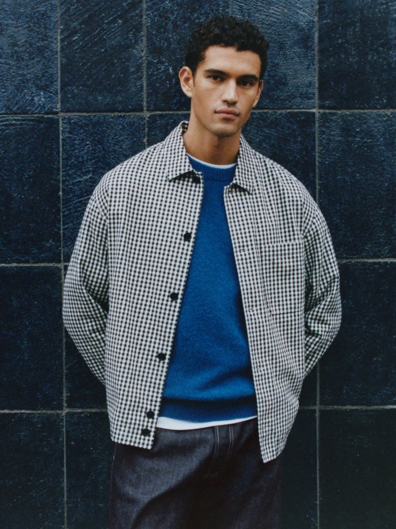 Portrait of a young man wearing a blue sweater and checkered jacket against a dark tiled wall, capturing a blend of casual fashion.