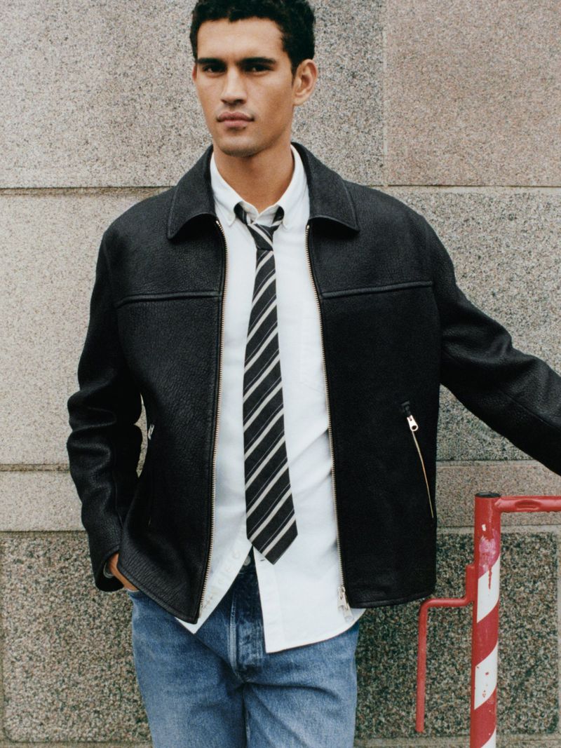 Portrait of a young adult man wearing a black jacket, white shirt, and tie, posing against a textured wall in an outdoor setting.