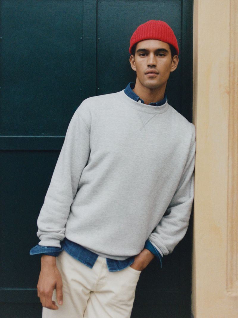 Portrait of an adult male wearing a light grey sweatshirt, cream pants, and a red knit beanie, leaning against a door outside.