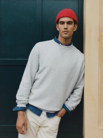Portrait of an adult male wearing a light grey sweatshirt, cream pants, and a red knit beanie, leaning against a door outside.