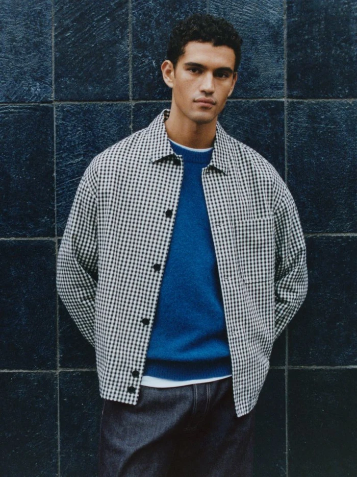 Model in a black and white check shirt over a blue sweater against a dark tile wall for Arket Real Denim.