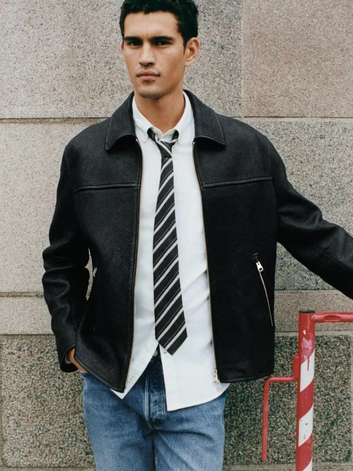 Model Bodhi Heeck wearing a black zip-up jacket, white collared shirt, striped tie, and blue denim jeans against a stone wall.