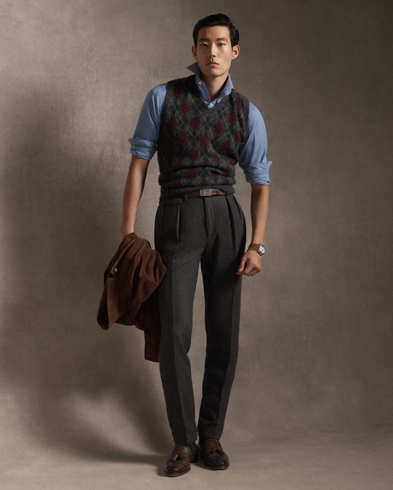 Full body shot of an Asian male model wearing a blue button-down shirt, argyle sweater vest, grey pleated pants, and brown loafers in studio.