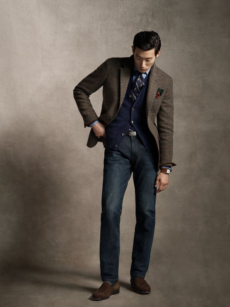 An Asian adult male is captured in a full-body studio shot, modelling a brown tweed blazer, navy waistcoat, jeans, and brown suede shoes.