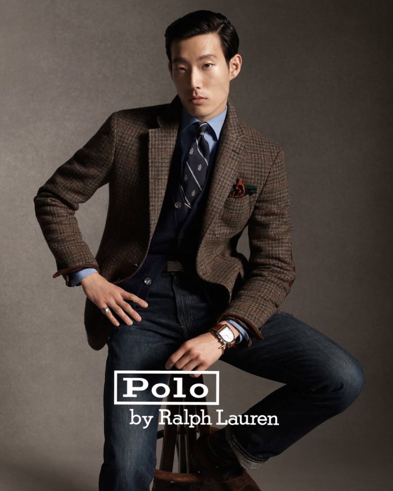 Man in brown tweed blazer, blue shirt, jeans, seated on stool. Ralph Lauren Polo branding visible. Fashion advertisement portrait.