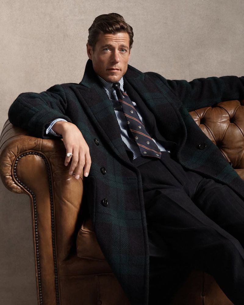 An adult male wearing a plaid overcoat, patterned tie, and striped shirt sits on a leather tufted couch in a brightly lit indoor setting.
