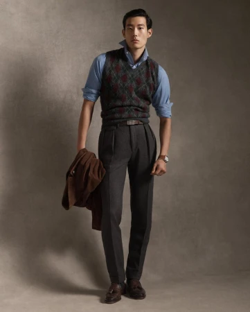 Model Juhyung Kang in an argyle sweater vest, blue shirt, and charcoal trousers holding a brown jacket against a textured studio backdrop.