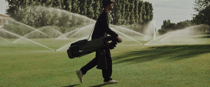 An adult male walks on a golf course, carrying a black golf bag, as sprinklers water the green grass and trees in the background.