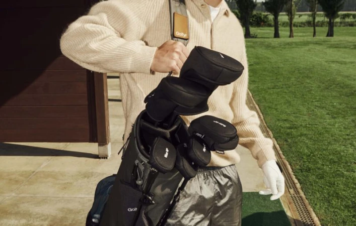 Person carrying a golf bag on a green golf course on a sunny day. The golfer is wearing a cream sweater and silver shorts.