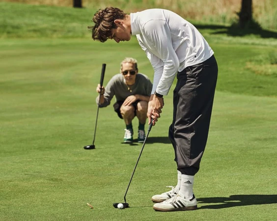 Two men play golf on a green course. One man tees up the ball while the other watches with a golf club in hand.