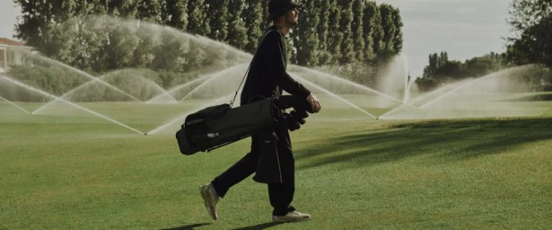Golfer in black attire carrying a golf bag across a green course with water sprinklers spraying in the background.