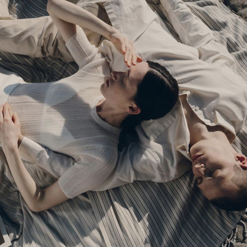 Overhead shot of two people lying head-to-head on a striped blanket outdoors, wearing neutral colored tops and pants in a minimalist style.