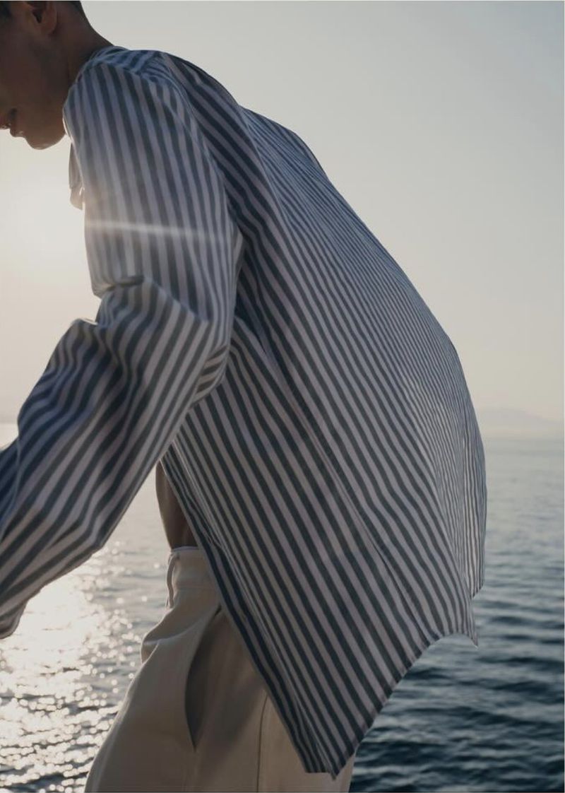 Man in a striped shirt stands near the ocean on a sunny day, showcasing a relaxed and stylish fashion aesthetic in an outdoor setting.