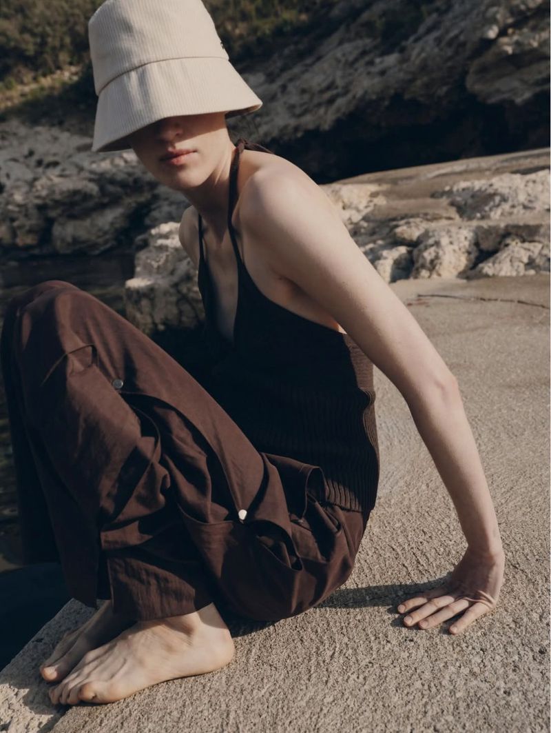 Woman in brown tank top and pants sits on concrete with a beige bucket hat in a bright outdoor setting near rocks.