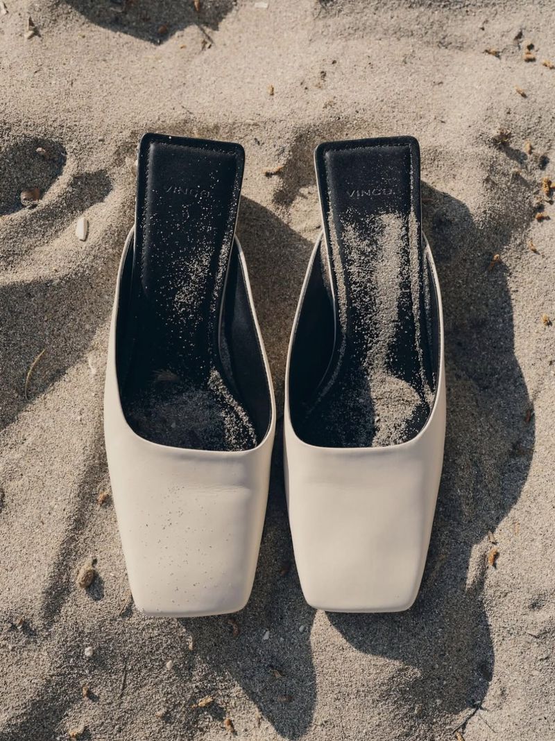 Two beige VINCE brand heeled mule shoes on tan sand in a high-angle shot with outdoor lighting, evoking fashion and summertime.