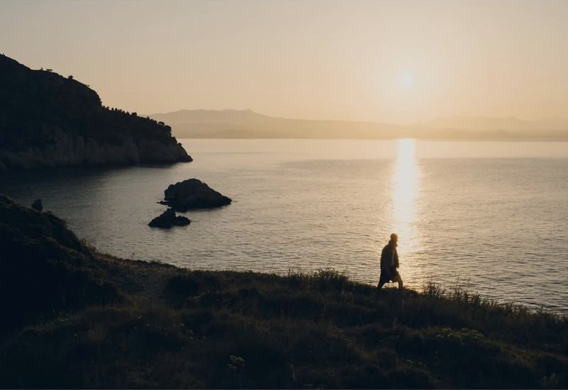 Person walking on a grassy hill overlooking the ocean at sunset, with mountains in the background, creating a peaceful outdoor scene.