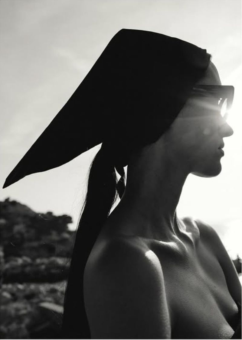 Monochrome portrait of a topless woman wearing sunglasses and a headscarf standing outdoors with a sky and rocky landscape background.