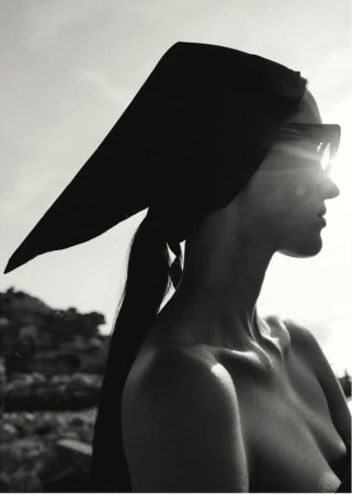 Monochrome portrait of a topless woman wearing sunglasses and a headscarf standing outdoors with a sky and rocky landscape background.