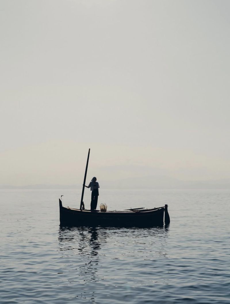 Silhouette of a person standing on a small fishing boat in calm, open water under a hazy sky; boat reflects in the blue water.