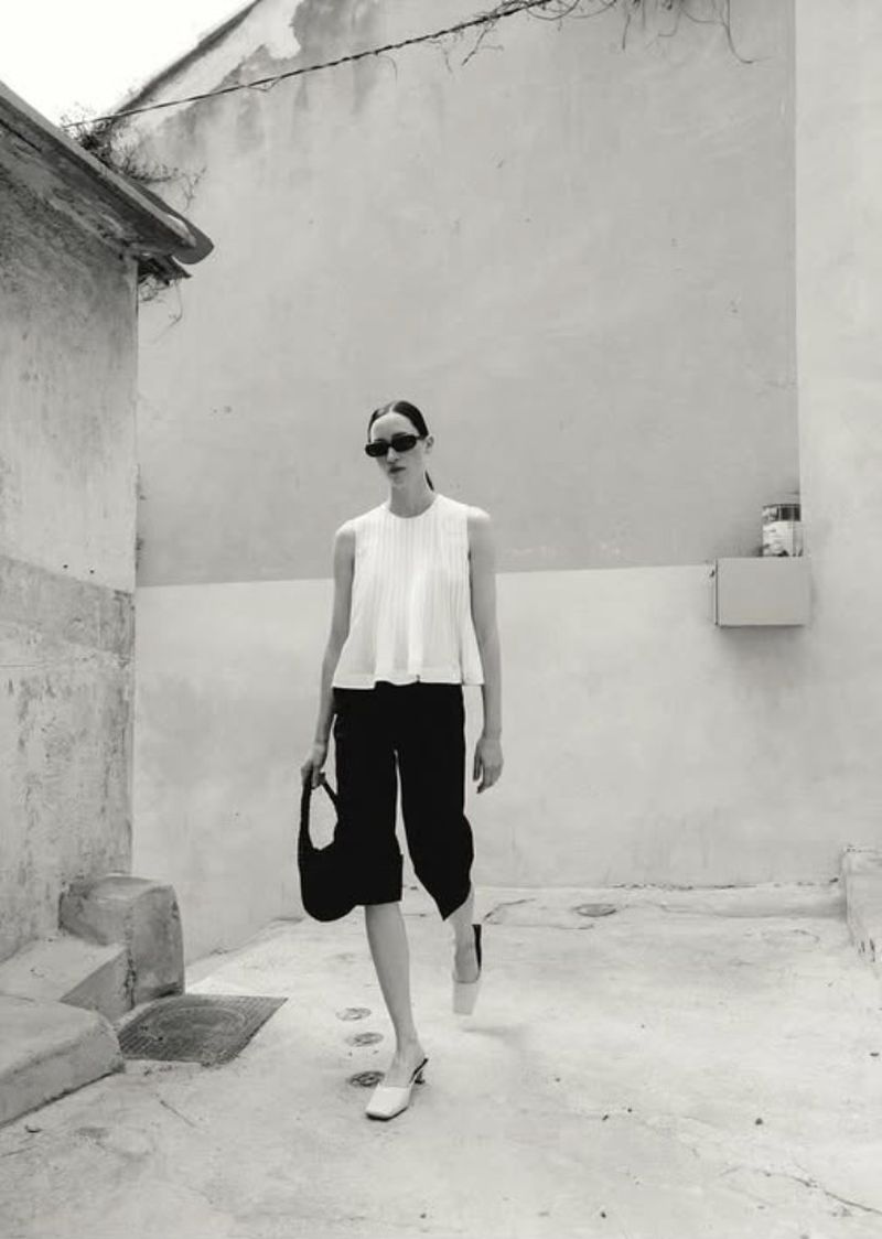 Monochrome full shot: woman in white top, black culottes, sunglasses, heels, and bag in sunlit alleyway. Fashion editorial style.
