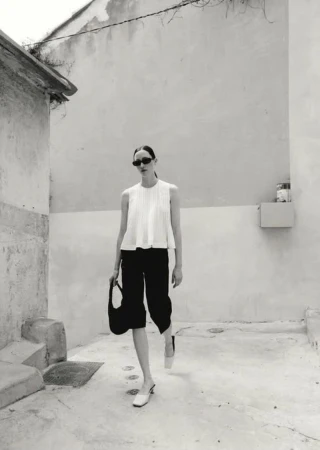 Monochrome full shot: woman in white top, black culottes, sunglasses, heels, and bag in sunlit alleyway. Fashion editorial style.