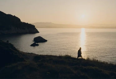 Model silhouetted walking along a grassy coastal cliff overlooking the calm ocean during a warm golden sunset.