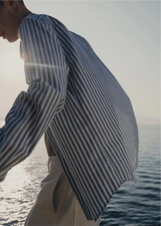 Man in a blue and white striped long-sleeve shirt and light trousers standing outdoors against the ocean sunlight.