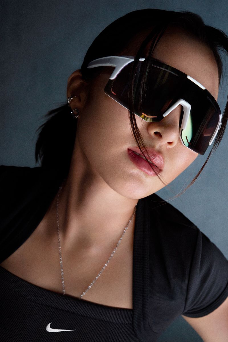 Portrait of a woman wearing white framed sunglasses, black shirt, and silver chain necklace in front of a grey backdrop.