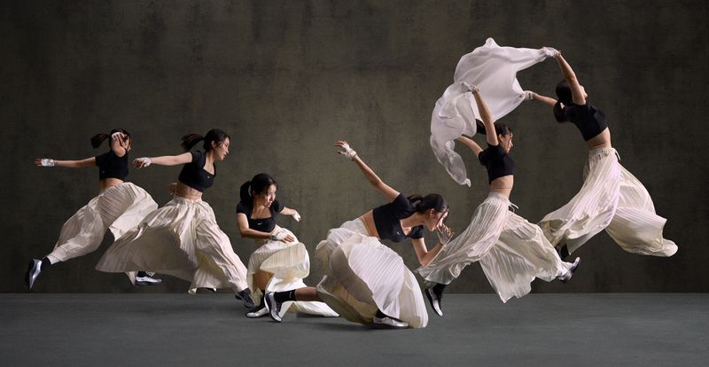 A woman in a black crop top and white pleated pants dynamically poses in multiple, overlapping frames against a gray background.