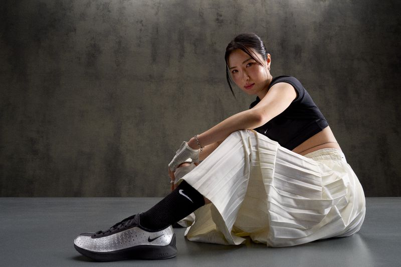 Asian woman poses seated in a photo studio wearing a black crop top, white pleated skirt, black socks, and silver Nike sneakers.
