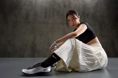 East Asian woman seated wearing a black crop top and white pleated skirt with metallic sneakers and black socks against a textured concrete studio background