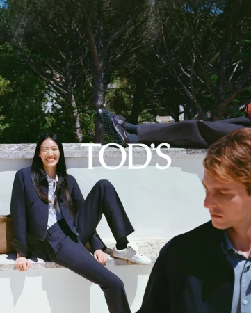 Fashion editorial featuring a smiling Asian woman in a navy suit, seated on a ledge, with a Caucasian man and greenery in the background.