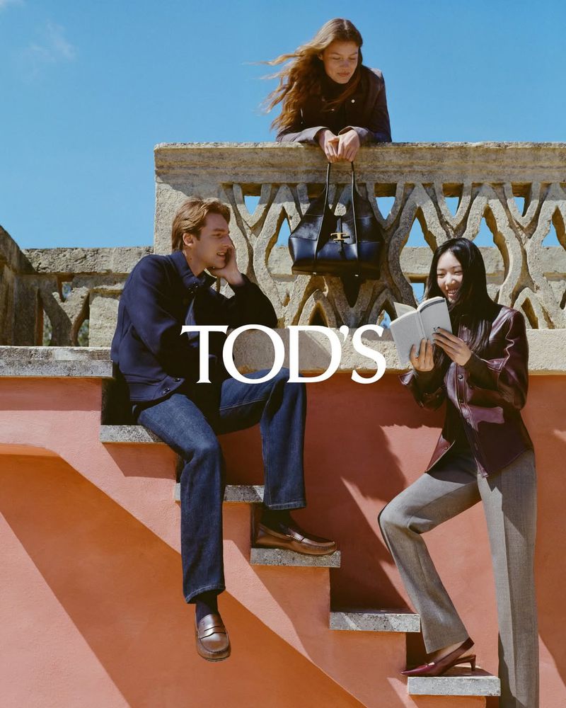 Three models posing on a terracotta staircase with an ornate carved stone balustrade and a black leather handbag for Tod's.