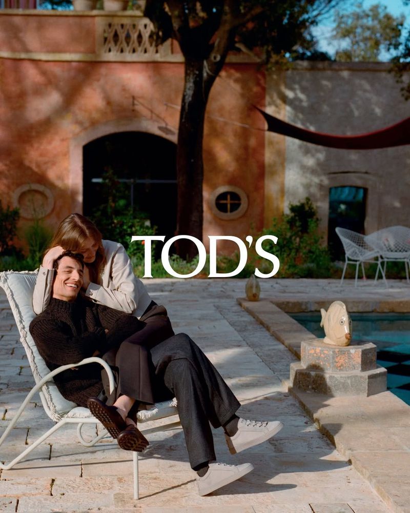Couple sitting on a chair outdoors, wearing casual clothing and Tod's shoes, near a pool and textured building, Tod's brand visible.