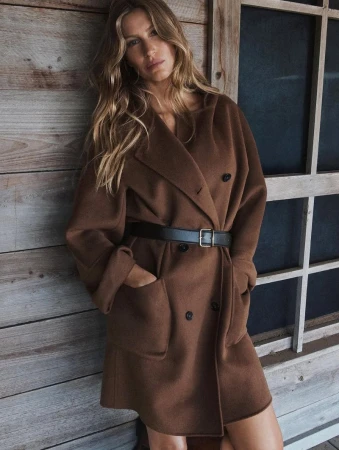 Adult woman with long hair posing in brown coat and black belt, with her hands in her pockets, leaning against wooden wall indoors.