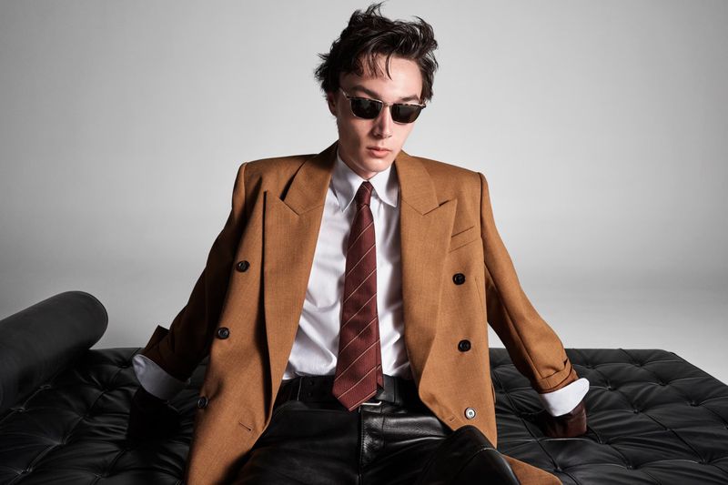 An adult male in a caramel double-breasted blazer, white collared shirt, black leather pants, and sunglasses sits on a black leather couch.