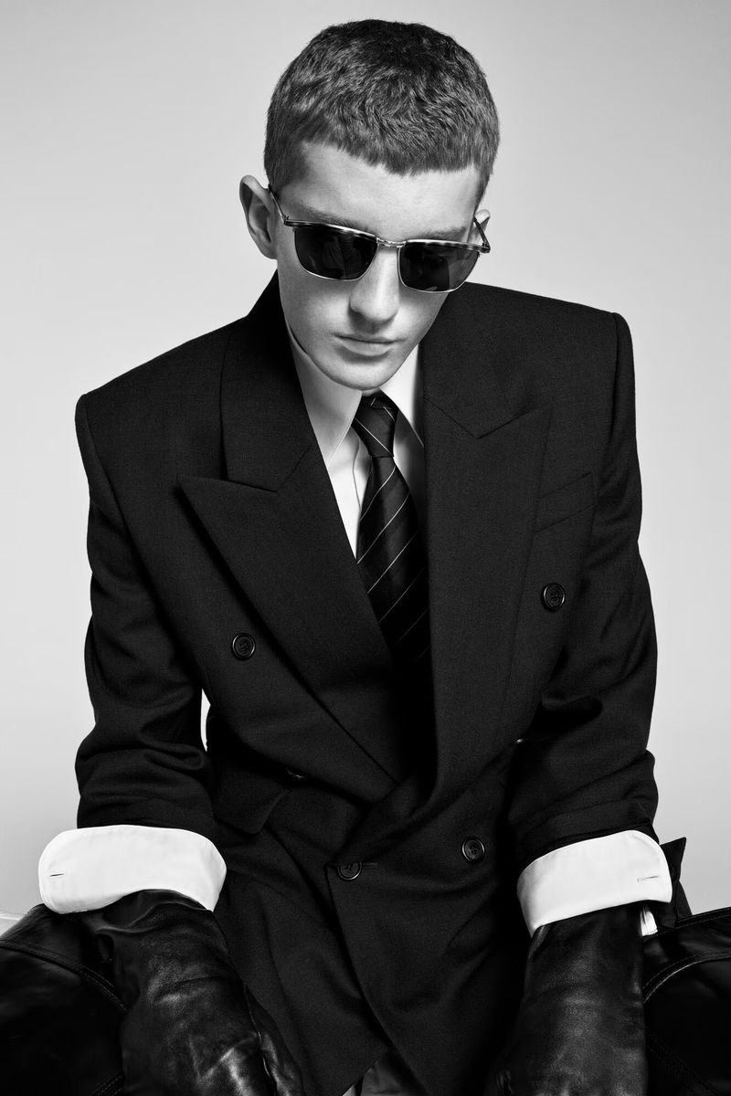 Monochrome portrait of a person in a black double-breasted suit, white shirt, and tie, accessorized with reflective sunglasses, indoors.