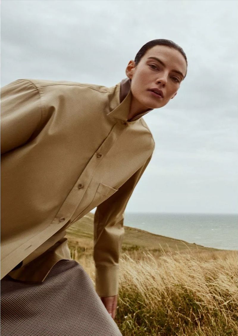 Adult female model in a tan button-down shirt poses on a grassy hill overlooking the sea under an overcast sky in a low-angle shot.