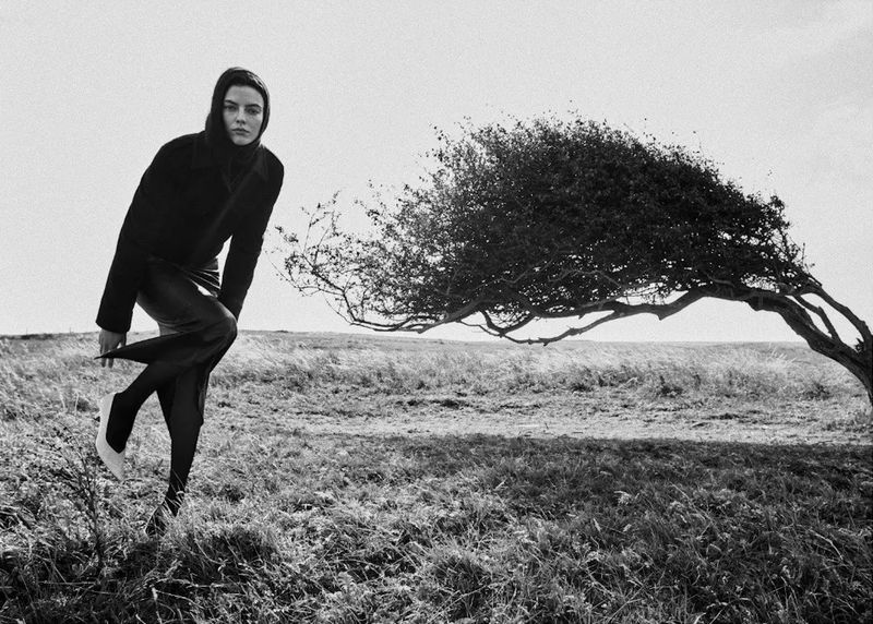 A woman in a black coat and dress poses in a grassy field with a windswept tree in a black and white fashion photograph on a sunny day.